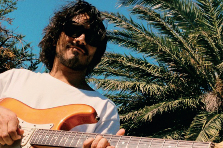 Musician Josh Morphett wearing sunglasses plays an electric guitar outdoors under a sunny blue sky with palm trees in the background.